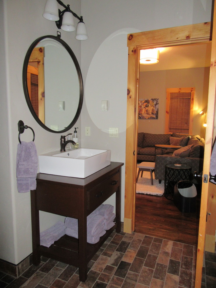Rustik inredning av ett litet badrum med dusch, med bruna skåp, en dusch i en alkov, en toalettstol med separat cisternkåpa, grå väggar, tegelgolv, ett fristående handfat, träbänkskiva, flerfärgat golv och dusch med gångjärnsdörr