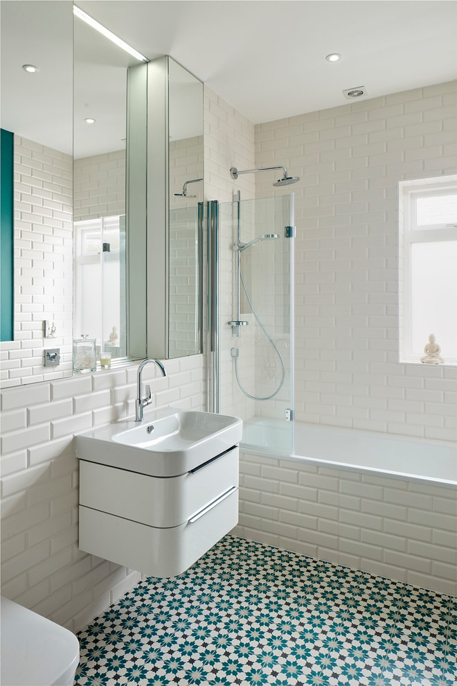 Photo of a contemporary bathroom in London with flat-panel cabinets, white cabinets, a built-in bath, a shower/bath combination, a wall mounted toilet, white tiles, metro tiles, white walls, a wall-mounted sink and multi-coloured floors.