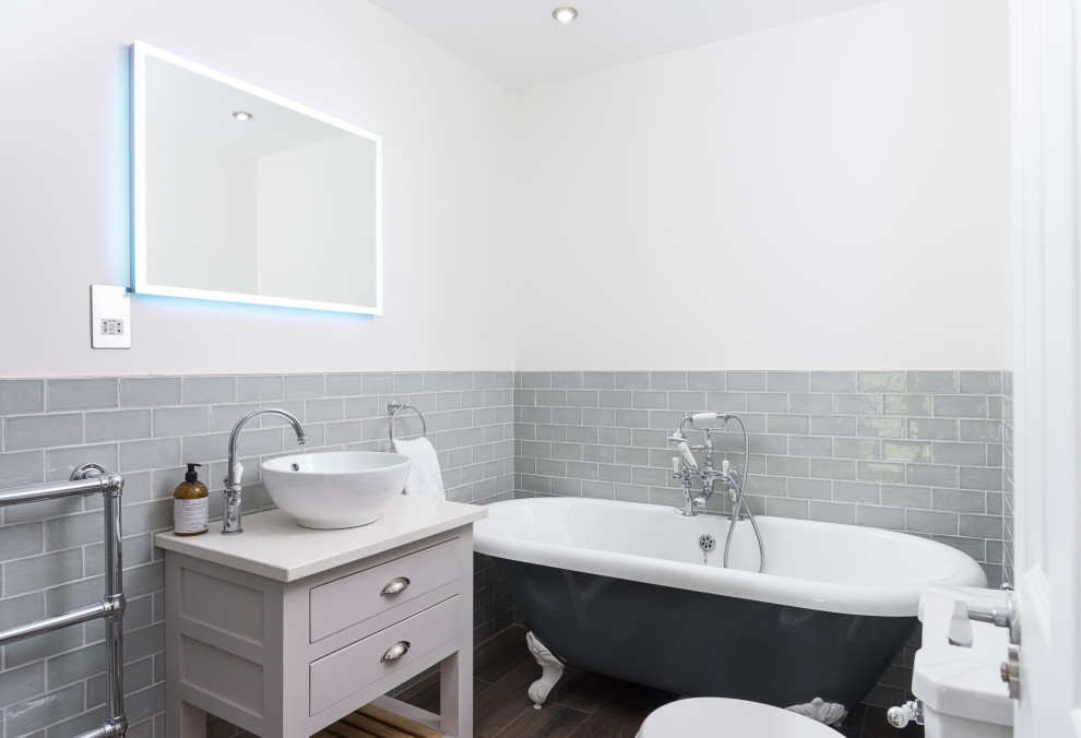 На фото: ванная комната в стиле неоклассика (современная классика) с плоскими фасадами, серыми фасадами, ванной на ножках, серой плиткой, плиткой кабанчик, белыми стенами, настольной раковиной, коричневым полом, белой столешницей и зеркалом с подсветкой
