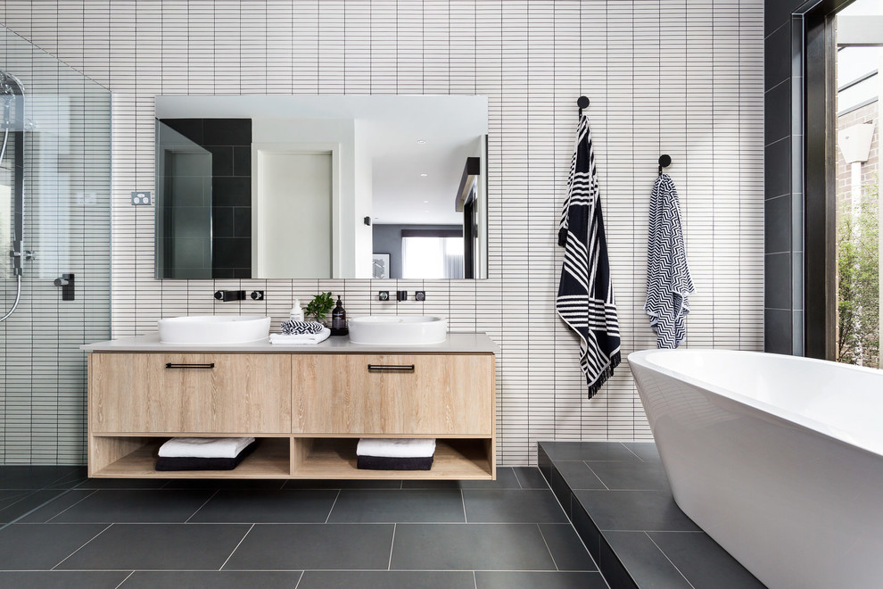 Modernes Badezimmer mit freistehender Badewanne, grauem Boden, flächenbündigen Schrankfronten, hellen Holzschränken, weißen Fliesen, Aufsatzwaschbecken und grauer Waschtischplatte in Melbourne