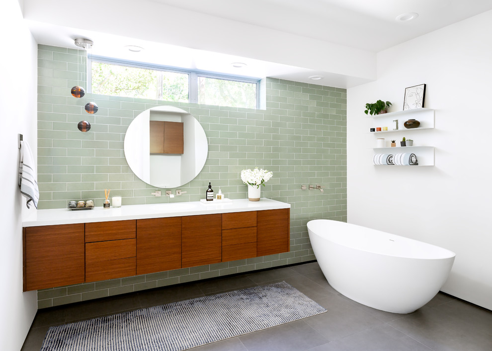 Bild på ett funkis vit vitt badrum med dusch, med släta luckor, skåp i mellenmörkt trä, ett fristående badkar, grön kakel, tunnelbanekakel, vita väggar, ett undermonterad handfat och grått golv