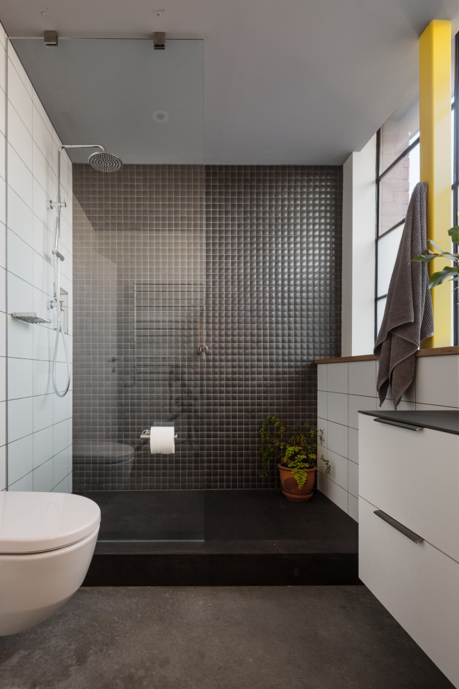 Inredning av ett industriellt svart svart badrum, med släta luckor, vita skåp, en dusch i en alkov, en vägghängd toalettstol, svart och vit kakel, betonggolv, grått golv och med dusch som är öppen