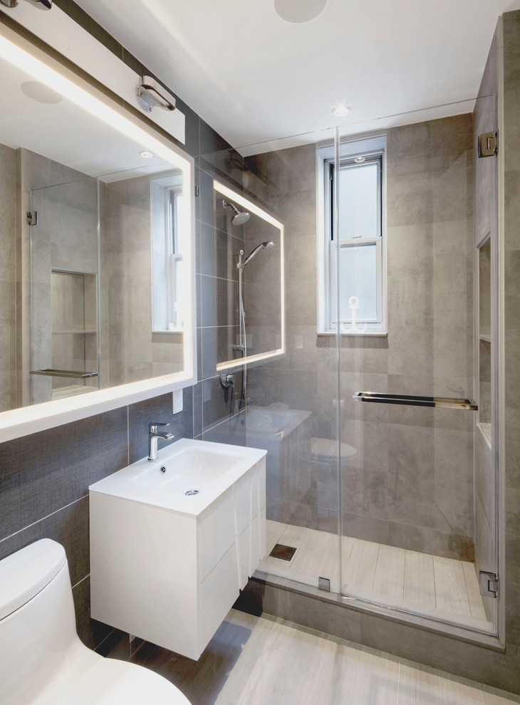 This is an example of a small contemporary ensuite bathroom in New York with freestanding cabinets, white cabinets, an alcove shower, a one-piece toilet, grey tiles, porcelain tiles, grey walls, porcelain flooring, a wall-mounted sink and glass worktops.