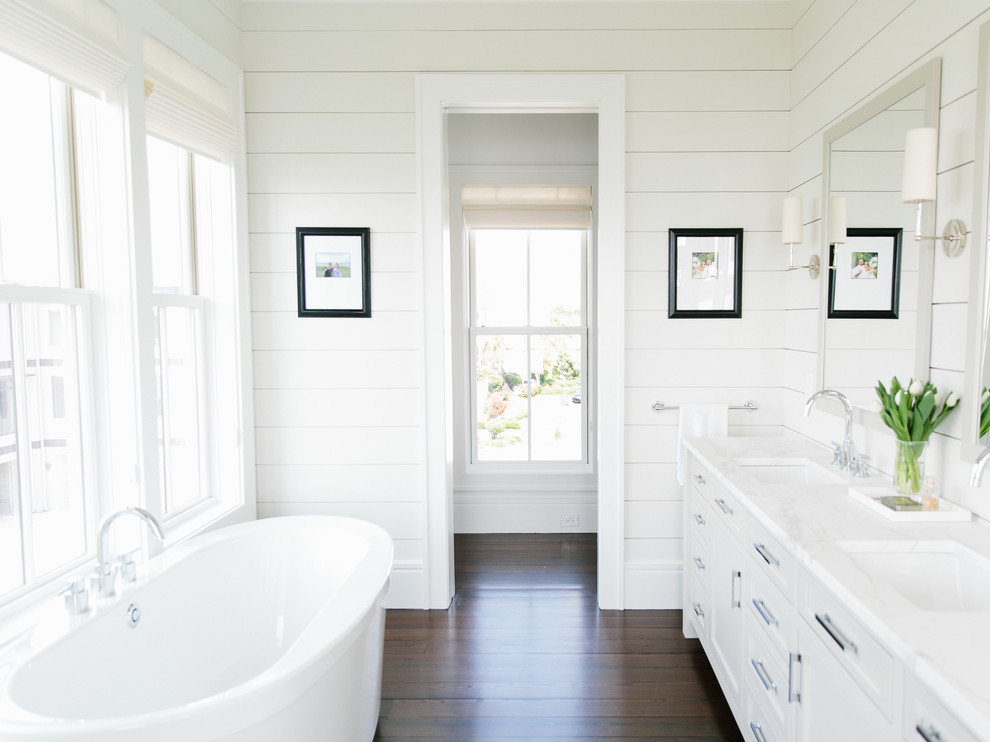 Exempel på ett klassiskt badrum, med luckor med infälld panel, vita skåp, ett fristående badkar, vita väggar, mörkt trägolv och ett undermonterad handfat