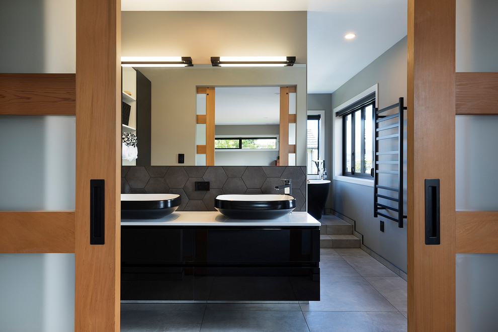 Réalisation d'une salle de bain principale design avec un placard à porte plane, des portes de placard noires, une baignoire indépendante, un carrelage gris, un mur gris, une vasque, un sol gris et un plan de toilette blanc.