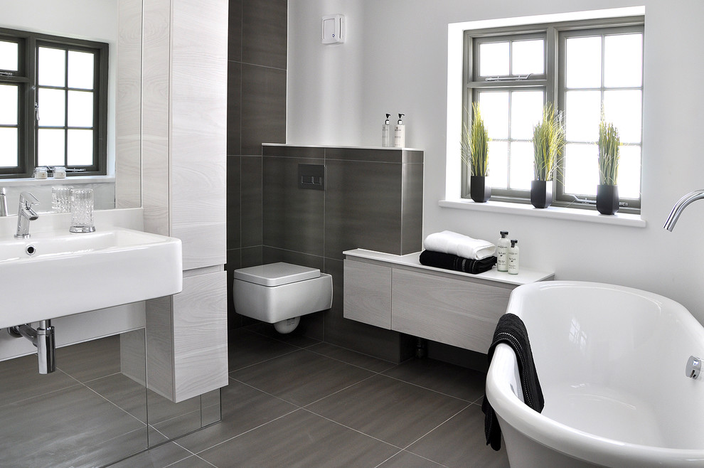 Modern inredning av ett badrum, med ett väggmonterat handfat, släta luckor, grå skåp, ett fristående badkar, en toalettstol med hel cisternkåpa, stenkakel, vita väggar och skiffergolv