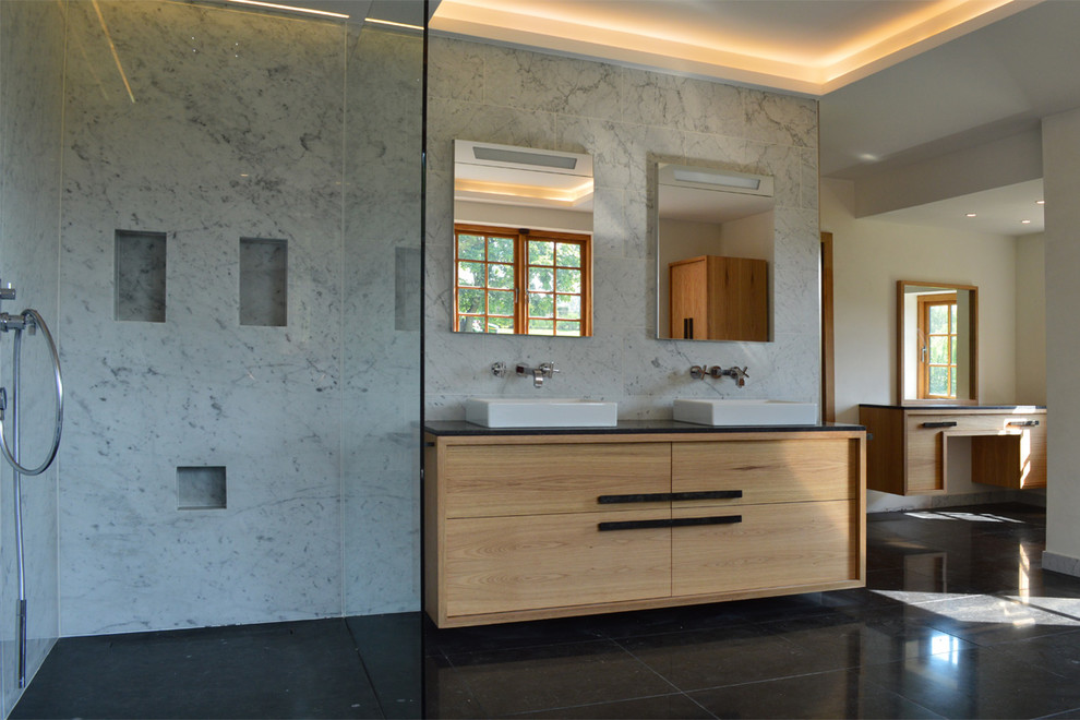Example of a large trendy master marble tile travertine floor and black floor bathroom design in Surrey with flat-panel cabinets, light wood cabinets, a wall-mount toilet and a drop-in sink