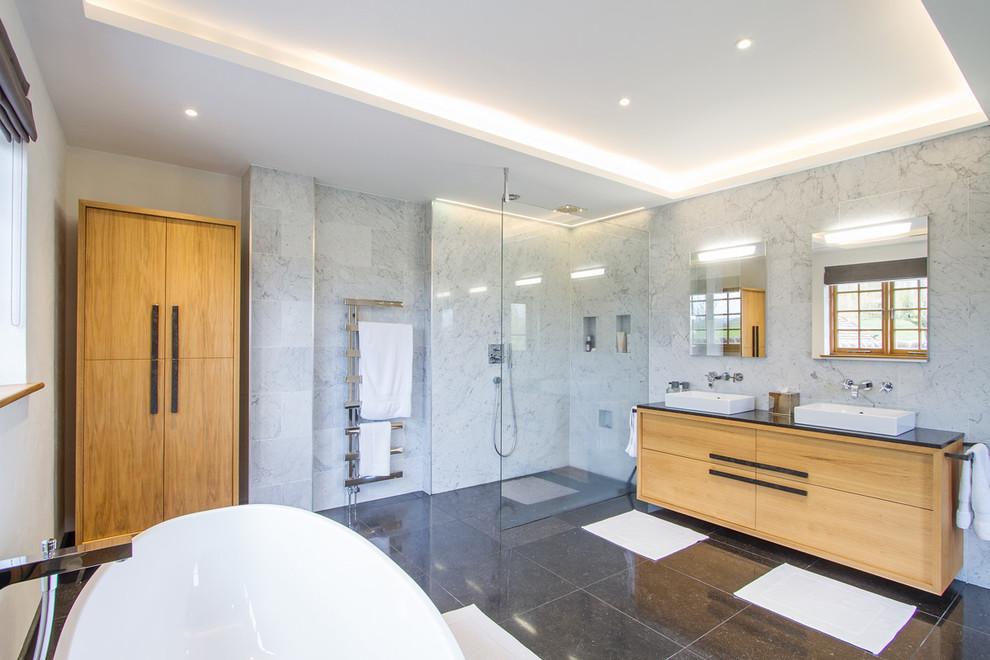Large trendy master marble tile travertine floor and black floor bathroom photo in Surrey with flat-panel cabinets, light wood cabinets, a wall-mount toilet and a drop-in sink