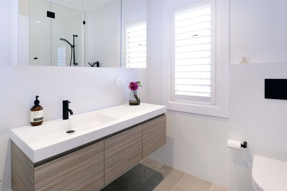 Пример оригинального дизайна: ванная комната среднего размера в современном стиле с плоскими фасадами, коричневыми фасадами, душем над ванной, раздельным унитазом, белой плиткой, керамической плиткой, белыми стенами, полом из керамической плитки, душевой кабиной, подвесной раковиной, столешницей из искусственного камня, серым полом, душем с распашными дверями и белой столешницей