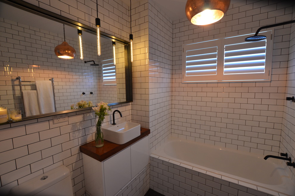 На фото: ванная комната в современном стиле с настольной раковиной, ванной в нише, душем над ванной, унитазом-моноблоком, плоскими фасадами, белыми фасадами, столешницей из дерева, белой плиткой и керамической плиткой