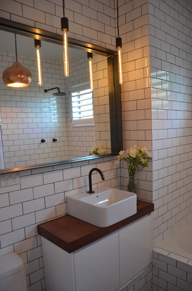 Пример оригинального дизайна: ванная комната в современном стиле с настольной раковиной, ванной в нише, душем над ванной, унитазом-моноблоком, плоскими фасадами, белыми фасадами, столешницей из дерева, белой плиткой и керамической плиткой