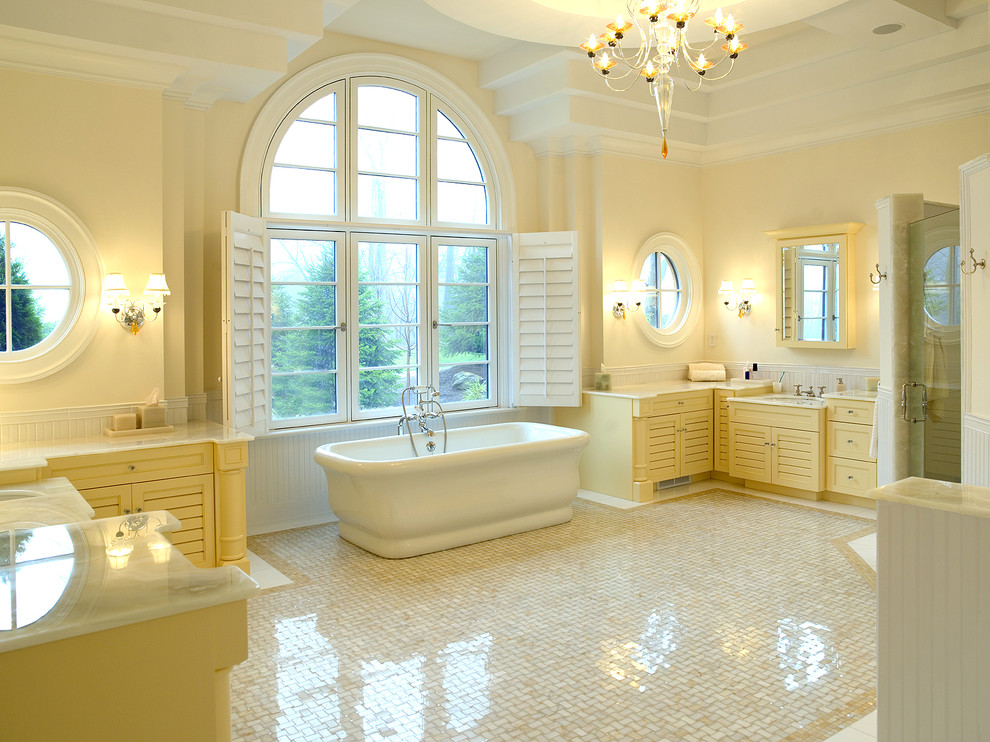 Идея дизайна: большая главная ванная комната в классическом стиле с фасадами с филенкой типа жалюзи, желтыми фасадами, отдельно стоящей ванной, угловым душем, белой плиткой, мраморной плиткой, бежевыми стенами, мраморным полом, врезной раковиной, столешницей из оникса, разноцветным полом и душем с распашными дверями