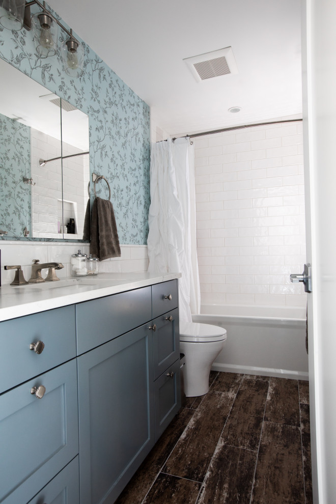 Small classic bathroom in Seattle with shaker cabinets, blue cabinets, an alcove bath, a shower/bath combination, a two-piece toilet, white tiles, metro tiles, blue walls, porcelain flooring, a submerged sink, engineered stone worktops, brown floors, a shower curtain, white worktops, a single sink and wallpapered walls.
