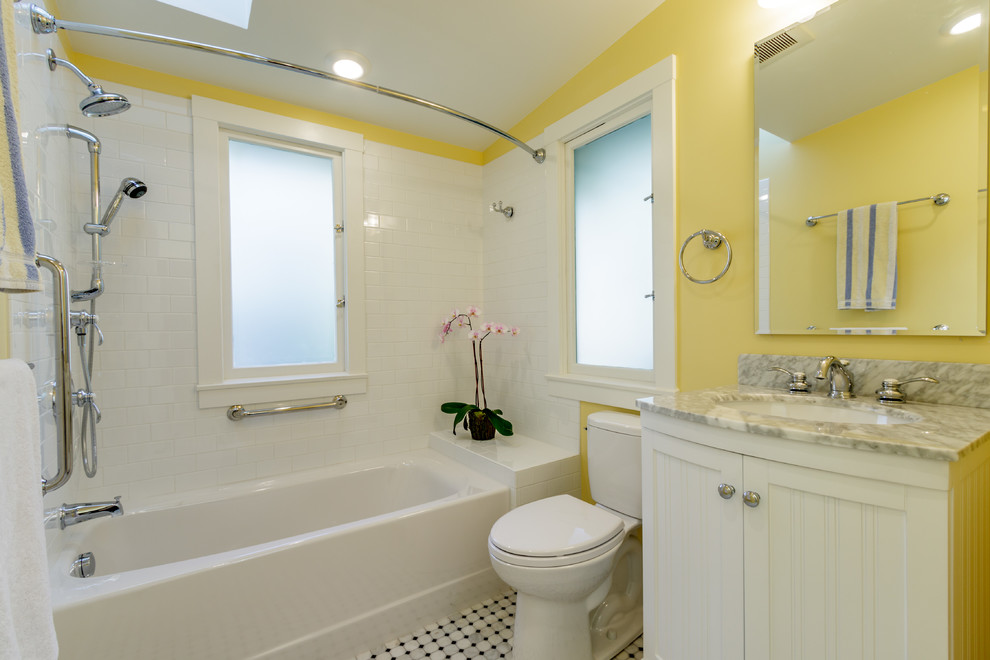 Amerikansk inredning av ett mellanstort badrum för barn, med ett undermonterad handfat, möbel-liknande, vita skåp, marmorbänkskiva, ett badkar i en alkov, en dusch/badkar-kombination, en toalettstol med separat cisternkåpa, vit kakel, keramikplattor, gula väggar och marmorgolv