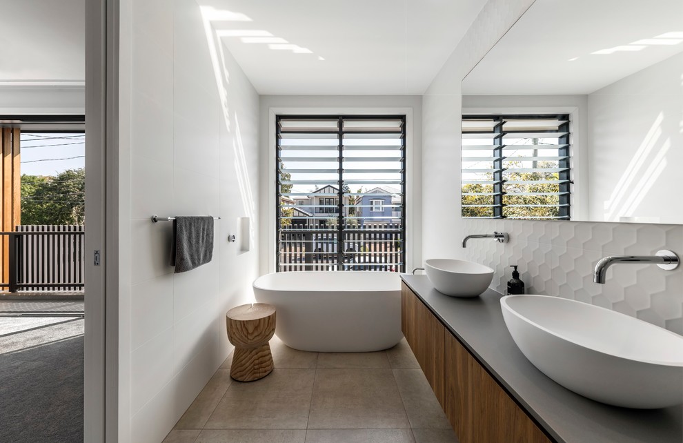 На фото: главная ванная комната в современном стиле с плоскими фасадами, фасадами цвета дерева среднего тона, отдельно стоящей ванной, белой плиткой, белыми стенами, настольной раковиной, бежевым полом и серой столешницей