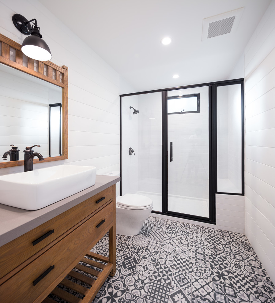 Пример оригинального дизайна: ванная комната среднего размера в стиле кантри с фасадами островного типа, светлыми деревянными фасадами, открытым душем, раздельным унитазом, белой плиткой, керамической плиткой, белыми стенами, полом из керамической плитки, душевой кабиной, настольной раковиной, столешницей из искусственного камня, разноцветным полом, душем с распашными дверями и серой столешницей