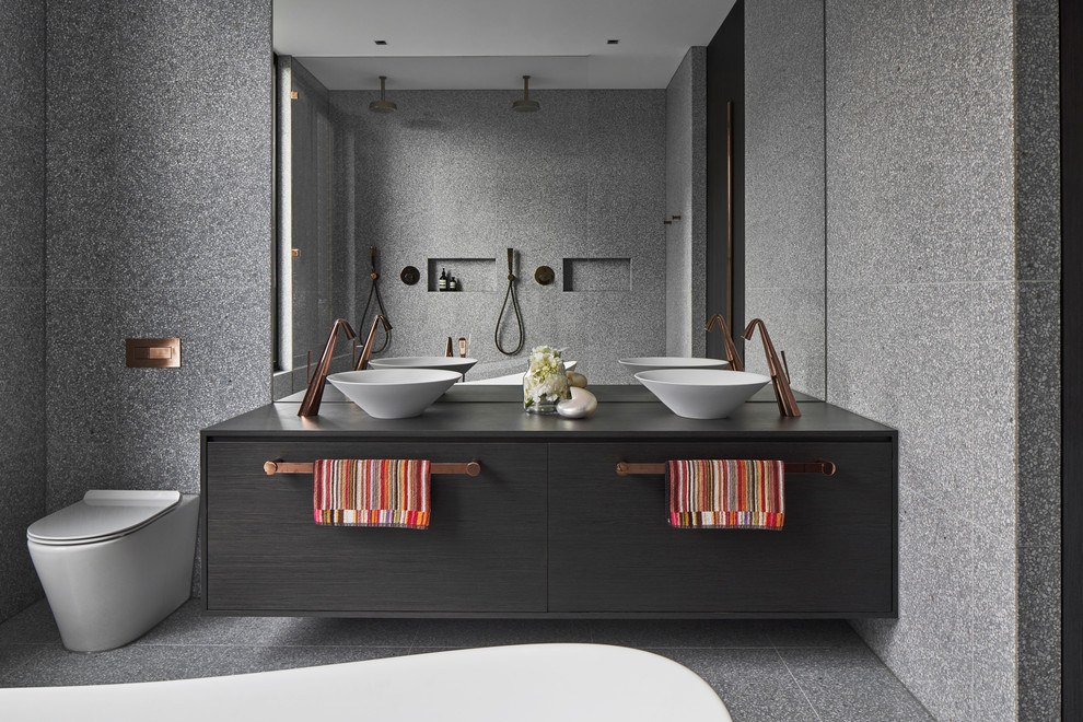 Modelo de cuarto de baño moderno con armarios con paneles lisos, puertas de armario negras, bañera exenta, baldosas y/o azulejos grises, lavabo sobreencimera, suelo gris y encimeras negras