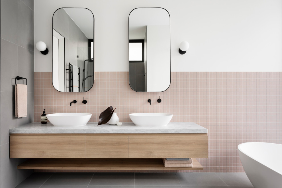 На фото: ванная комната в современном стиле с плоскими фасадами, светлыми деревянными фасадами, отдельно стоящей ванной, розовой плиткой, плиткой мозаикой, белыми стенами, настольной раковиной, серым полом, белой столешницей, тумбой под две раковины и подвесной тумбой
