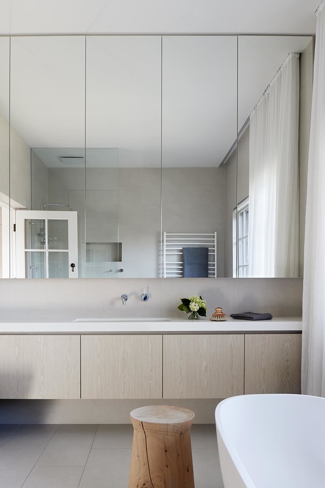 На фото: ванная комната в современном стиле с плоскими фасадами, светлыми деревянными фасадами, отдельно стоящей ванной, бежевой плиткой, бежевыми стенами, врезной раковиной, бежевым полом и белой столешницей