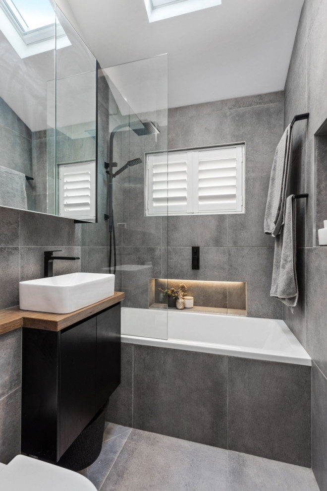 Inspiration for a small contemporary ensuite bathroom in Sydney with flat-panel cabinets, black cabinets, a built-in bath, a shower/bath combination, a wall mounted toilet, grey tiles, ceramic tiles, grey walls, ceramic flooring, a vessel sink, wooden worktops, grey floors, brown worktops, a wall niche, a single sink, a built in vanity unit and a vaulted ceiling.