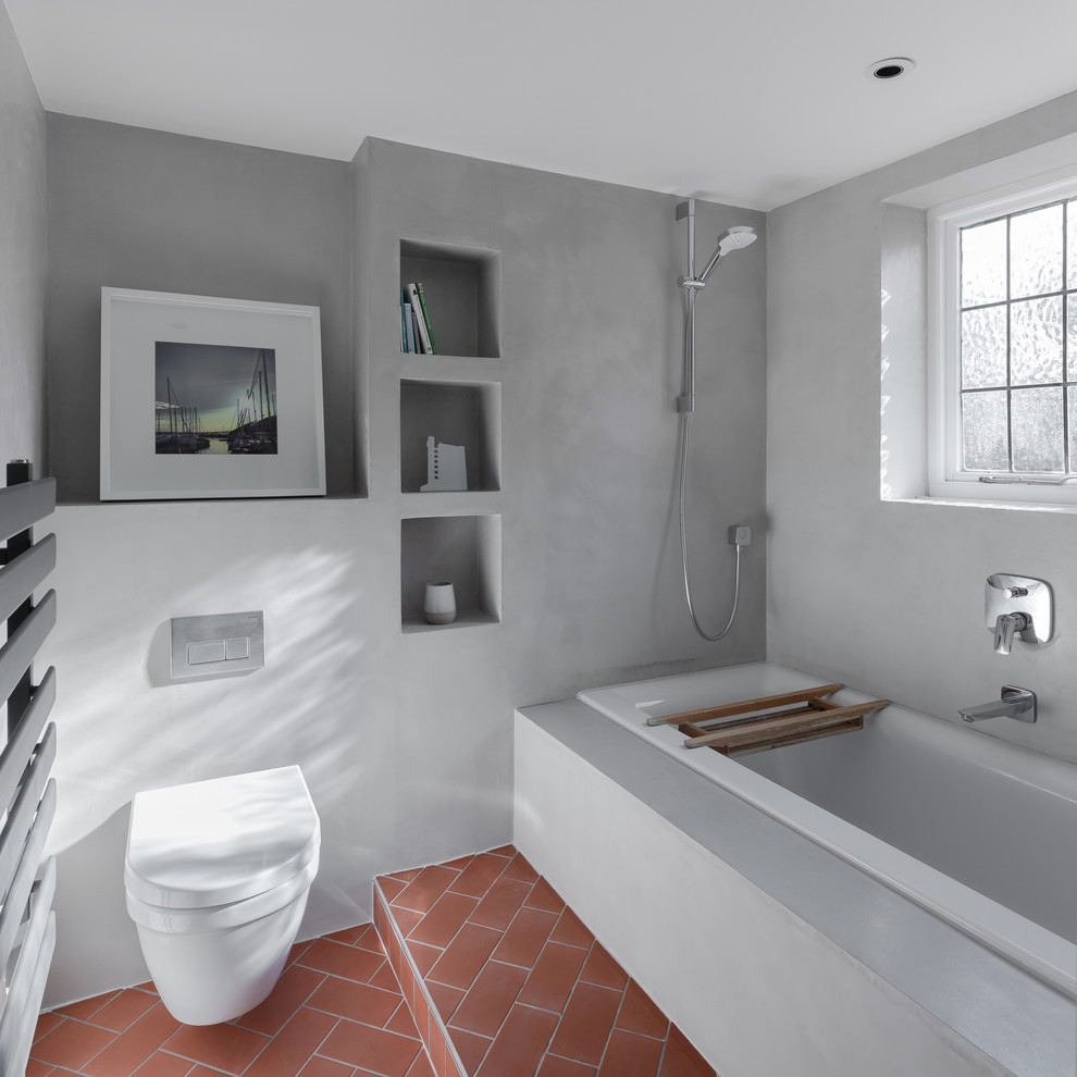 Exempel på ett mellanstort nordiskt beige beige badrum för barn, med ett platsbyggt badkar, en dusch/badkar-kombination, en vägghängd toalettstol, grå kakel, grå väggar, klinkergolv i keramik, ett avlångt handfat, träbänkskiva, rött golv och med dusch som är öppen