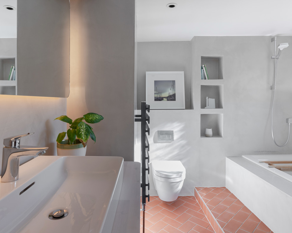 Photo of a medium sized scandi family bathroom in Oxfordshire with a built-in bath, a shower/bath combination, a wall mounted toilet, grey tiles, grey walls, ceramic flooring, a trough sink, wooden worktops, red floors, an open shower and beige worktops.