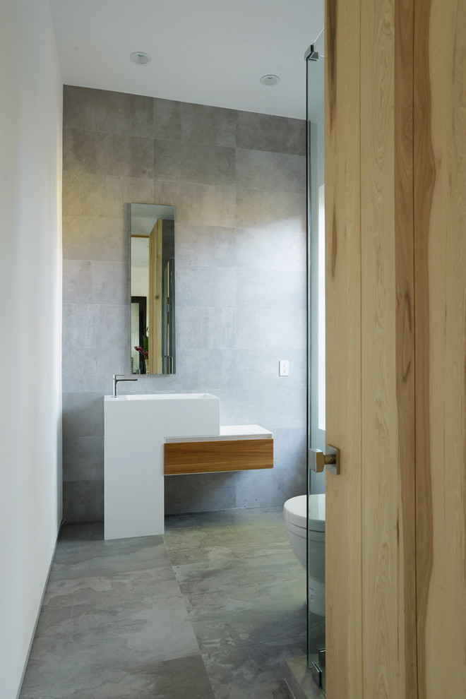 Inredning av ett modernt litet badrum med dusch, med ett piedestal handfat, grå kakel, cementkakel, betonggolv, släta luckor, skåp i ljust trä, en hörndusch, en toalettstol med separat cisternkåpa, grå väggar och bänkskiva i akrylsten