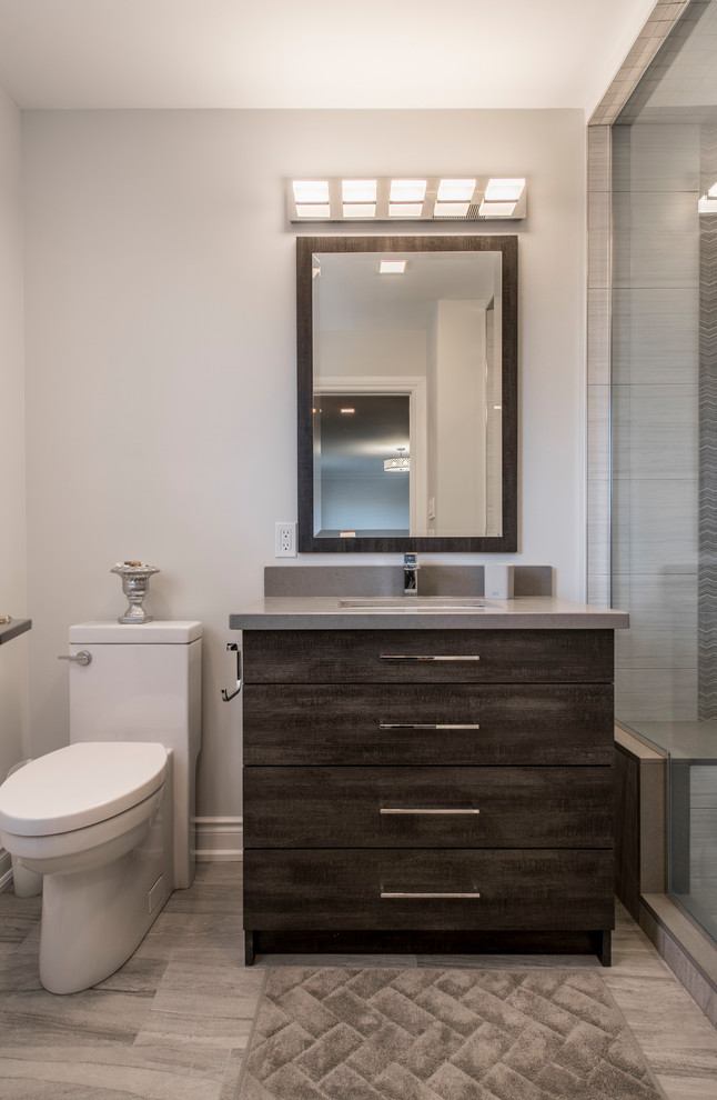 Exemple d'une salle de bain tendance en bois foncé de taille moyenne avec un placard à porte plane, un carrelage gris, des carreaux de porcelaine, un mur gris, un sol en vinyl, un lavabo encastré, un plan de toilette en surface solide, un sol gris, une cabine de douche à porte battante et un plan de toilette gris.