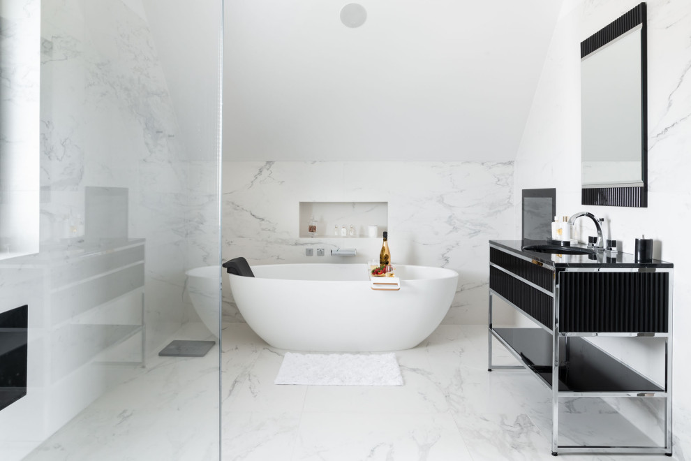 Expansive contemporary ensuite bathroom in London with freestanding cabinets, black cabinets, a freestanding bath, a walk-in shower, a wall mounted toilet, white tiles, marble tiles, white walls, marble flooring, a built-in sink, marble worktops, white floors, an open shower and black worktops.