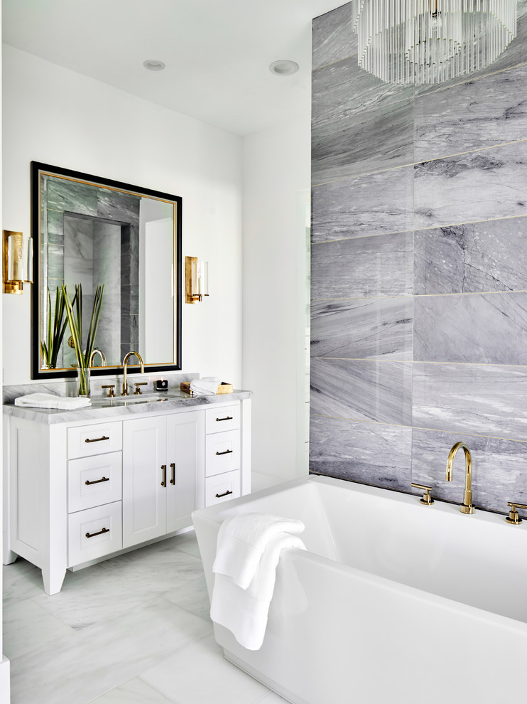 Cette photo montre une salle d'eau chic avec un placard à porte shaker, des portes de placard blanches, une baignoire indépendante, un carrelage gris, du carrelage en marbre, un mur blanc, un sol en marbre, un lavabo encastré, un plan de toilette en marbre, un sol blanc et un plan de toilette gris.