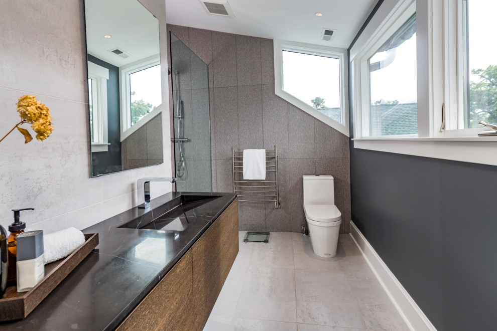 Example of a mid-sized trendy master gray tile and ceramic tile marble floor and white floor bathroom design in New York with flat-panel cabinets, medium tone wood cabinets, a one-piece toilet, white walls, a wall-mount sink, marble countertops and black countertops
