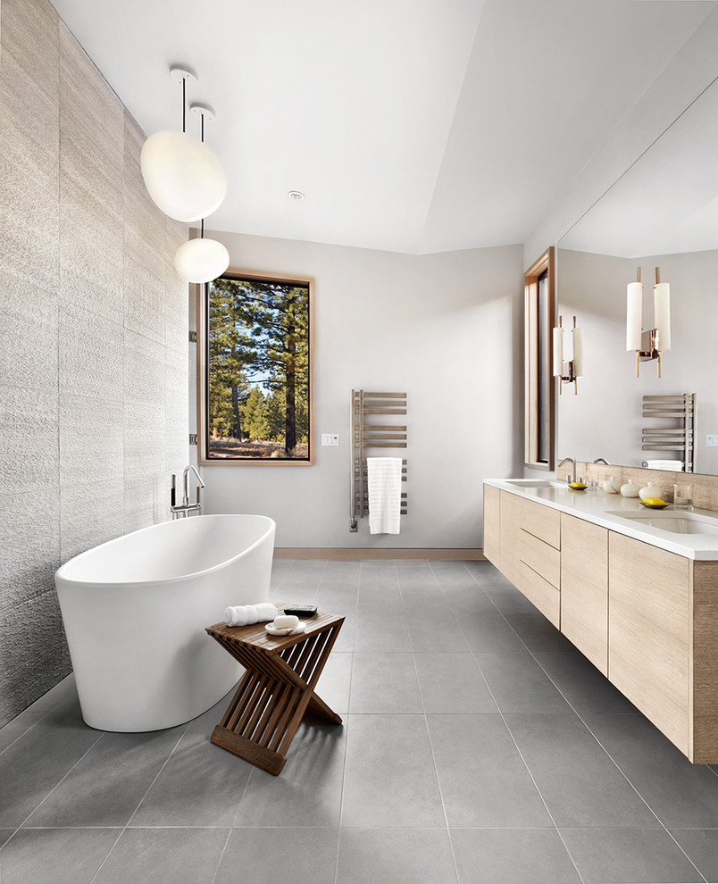 Inredning av ett modernt badrum, med ett undermonterad handfat, släta luckor, skåp i ljust trä, ett fristående badkar, grå kakel, grå väggar och grått golv