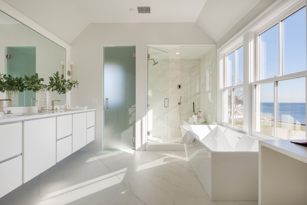 Inspiration for a large coastal ensuite bathroom in New York with flat-panel cabinets, white cabinets, a freestanding bath, an alcove shower, white tiles, stone slabs, porcelain flooring, a submerged sink, marble worktops, white floors, a hinged door, white worktops and grey walls.