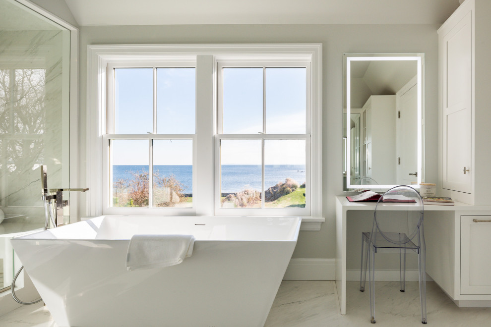 Inspiration for a large beach style ensuite bathroom in New York with flat-panel cabinets, white cabinets, a freestanding bath, an alcove shower, a one-piece toilet, white tiles, stone slabs, beige walls, porcelain flooring, a submerged sink, marble worktops, white floors, a hinged door and white worktops.