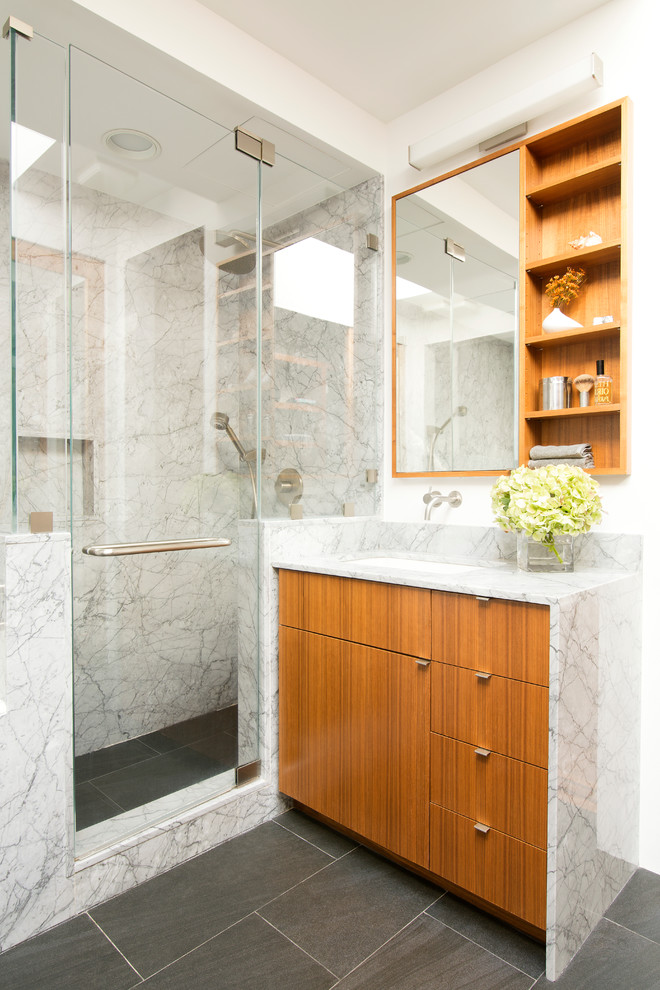 Medium sized contemporary bathroom in New York with flat-panel cabinets, medium wood cabinets, a corner shower, a one-piece toilet, grey tiles, stone slabs, white walls, porcelain flooring, a submerged sink, marble worktops and a hinged door.