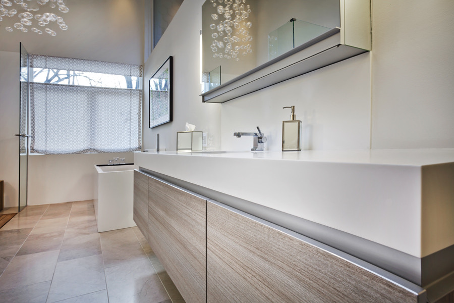 This is an example of a large modern ensuite bathroom in Minneapolis with a submerged sink, flat-panel cabinets, light wood cabinets, solid surface worktops, a freestanding bath, a built-in shower, a one-piece toilet, grey tiles, stone tiles, white walls and limestone flooring.