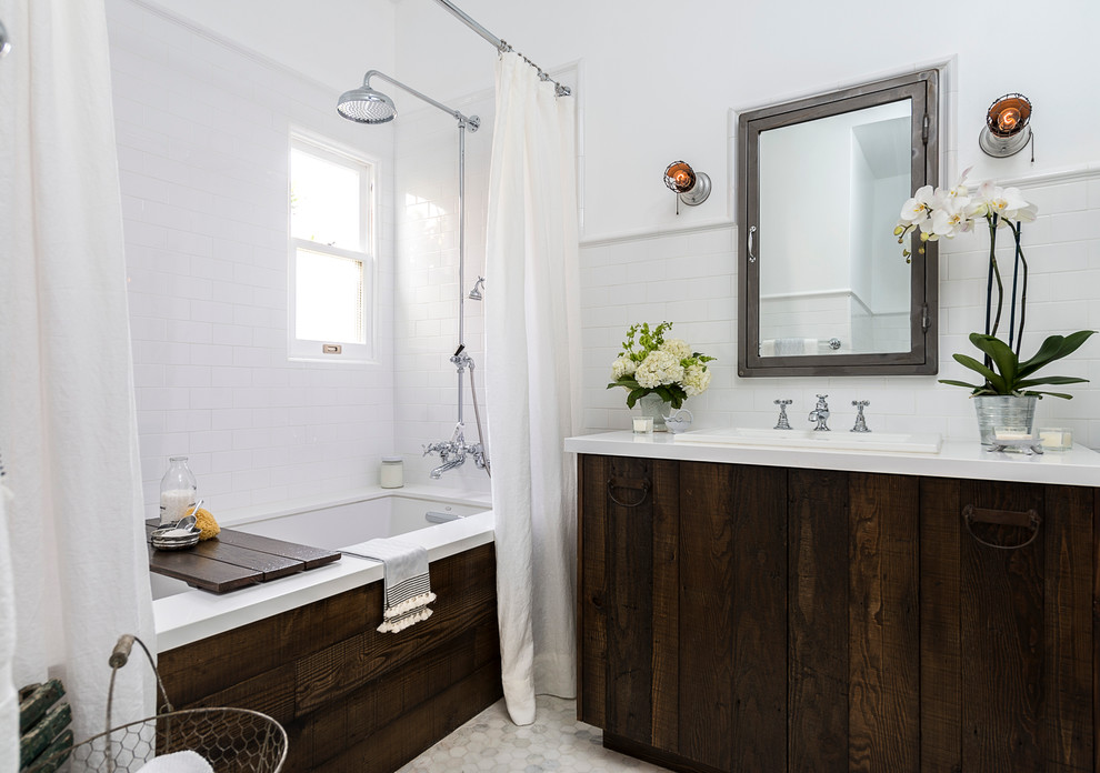 Inredning av ett lantligt litet en-suite badrum, med ett nedsänkt handfat, släta luckor, ett undermonterat badkar, en dusch/badkar-kombination, en toalettstol med separat cisternkåpa, vit kakel, tunnelbanekakel, vita väggar, marmorgolv, bänkskiva i akrylsten, skåp i slitet trä och dusch med duschdraperi