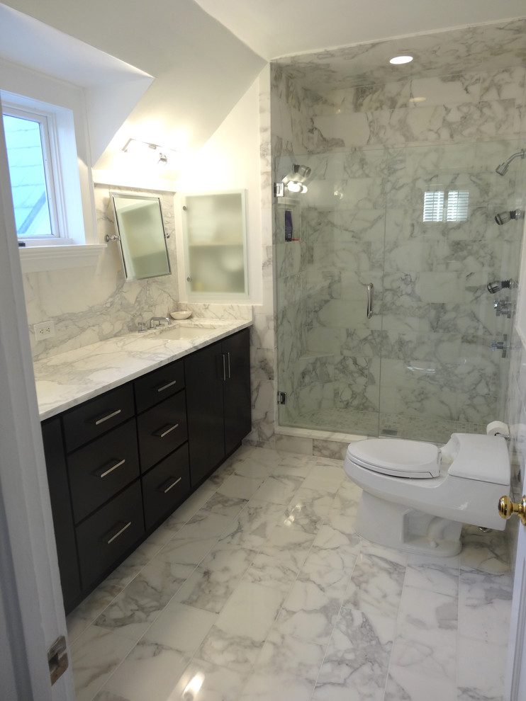 Modern inredning av ett badrum, med ett undermonterad handfat, släta luckor, skåp i mörkt trä, marmorbänkskiva, en toalettstol med hel cisternkåpa, vit kakel och stenkakel