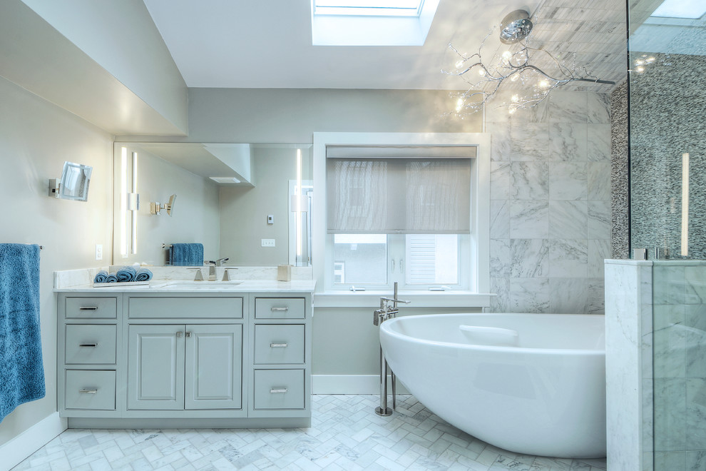 Cette image montre une salle de bain principale traditionnelle de taille moyenne avec des portes de placard grises, une baignoire indépendante, un carrelage blanc, un carrelage de pierre, un mur gris, un lavabo encastré, une douche d'angle et un placard avec porte à panneau surélevé.