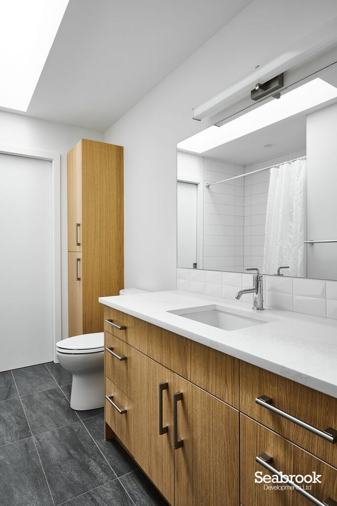 Bild på ett mellanstort funkis vit vitt en-suite badrum, med släta luckor, skåp i ljust trä, ett platsbyggt badkar, en dusch/badkar-kombination, en toalettstol med hel cisternkåpa, vit kakel, vita väggar, klinkergolv i porslin, ett undermonterad handfat, bänkskiva i kvarts, grått golv och dusch med duschdraperi