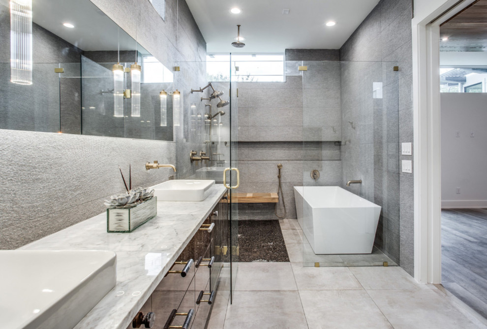 Photo of a large contemporary ensuite wet room bathroom in Dallas with flat-panel cabinets, brown cabinets, a freestanding bath, grey tiles, pebble tiles, grey walls, porcelain flooring, a built-in sink, marble worktops, grey floors, a hinged door and grey worktops.