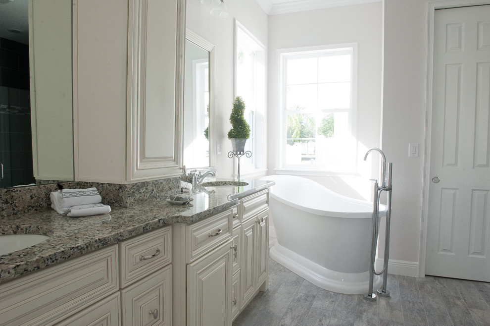 Design ideas for a farmhouse ensuite bathroom in Orlando with raised-panel cabinets, distressed cabinets, a freestanding bath, a built-in shower, grey tiles, porcelain tiles, grey walls, porcelain flooring, a submerged sink and granite worktops.