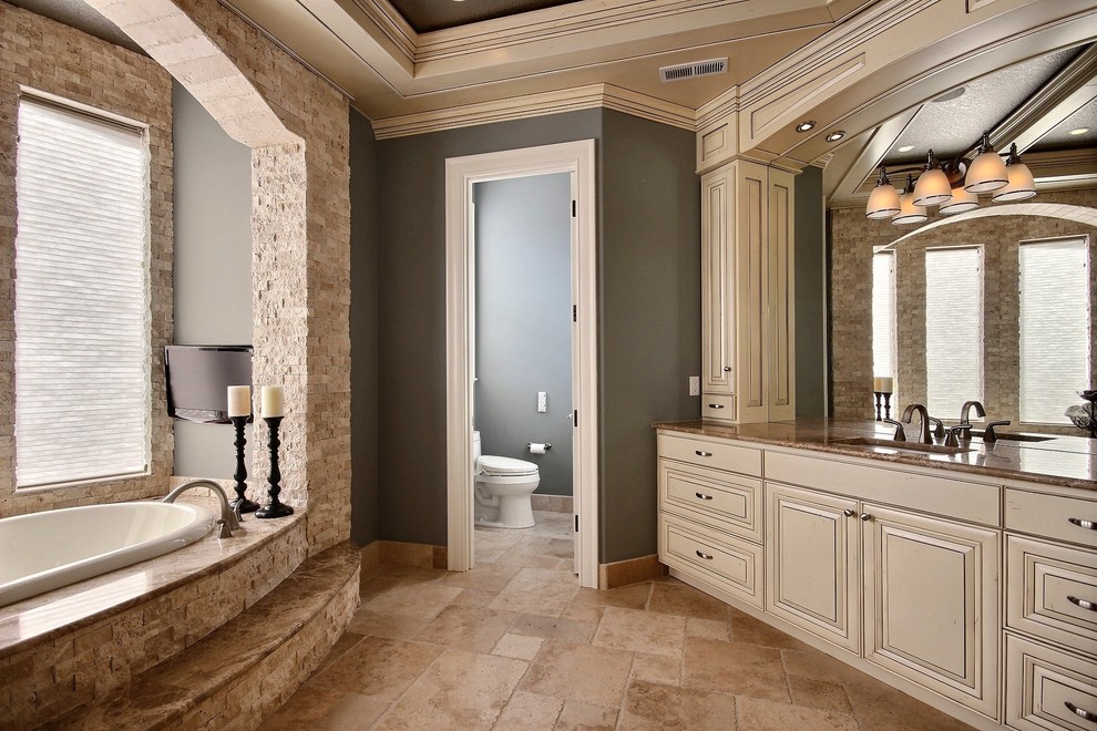 This is an example of an expansive classic ensuite bathroom in Portland with raised-panel cabinets, beige cabinets, an alcove bath, a one-piece toilet, beige tiles, stone slabs, blue walls, travertine flooring, a submerged sink and marble worktops.