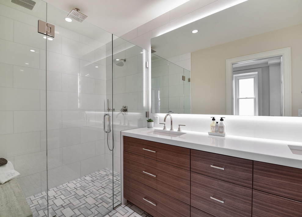This is an example of an ensuite bathroom in Boston with flat-panel cabinets, dark wood cabinets, a built-in shower, white tiles, white walls, mosaic tile flooring, a submerged sink, solid surface worktops, grey floors, a hinged door, a shower bench, double sinks and a built in vanity unit.