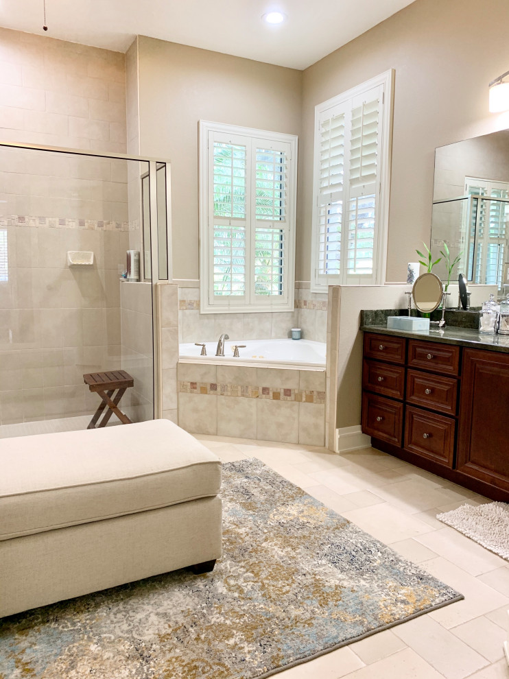 This is an example of a large traditional ensuite bathroom in Orlando with raised-panel cabinets, brown cabinets, a corner bath, an alcove shower, a one-piece toilet, beige tiles, ceramic tiles, grey walls, ceramic flooring, a submerged sink, granite worktops, beige floors, a hinged door, black worktops, a shower bench, double sinks and a built in vanity unit.