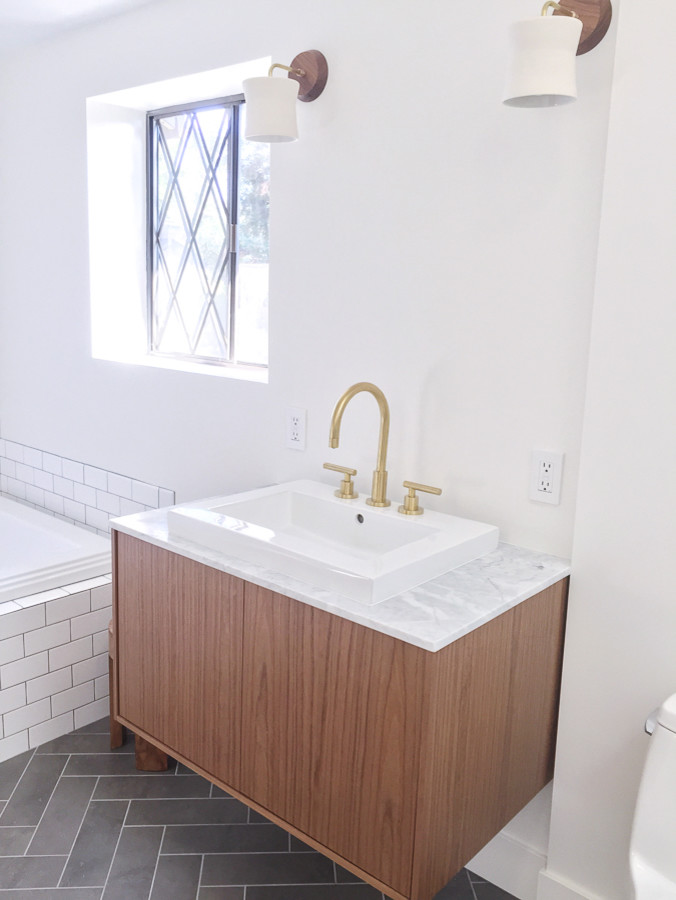 Modelo de cuarto de baño principal, de estilo de casa de campo, de tamaño medio, con armarios con paneles lisos, puertas de armario de madera oscura, bañera encastrada, ducha a ras de suelo, sanitario de una pieza, baldosas y/o azulejos blancos, baldosas y/o azulejos de cemento, paredes blancas, suelo de baldosas de porcelana, lavabo sobreencimera, encimera de mármol, suelo gris, ducha con puerta con bisagras y encimeras blancas