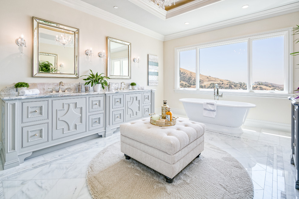 Modelo de cuarto de baño mediterráneo con armarios con rebordes decorativos, puertas de armario grises, bañera exenta, paredes beige, suelo blanco y encimeras grises