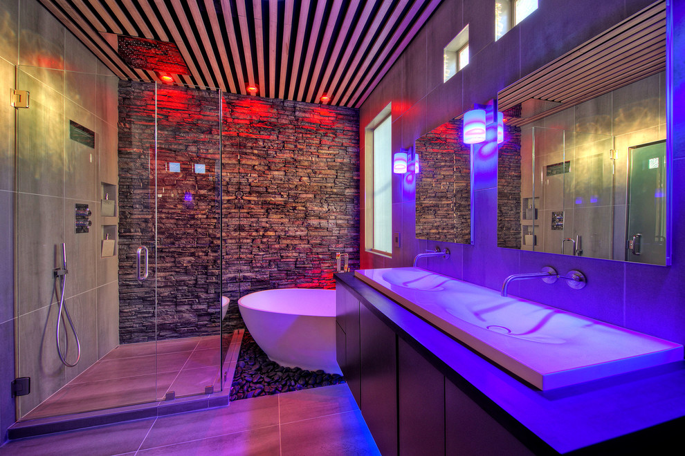 Exempel på ett mellanstort modernt en-suite badrum, med släta luckor, skåp i mörkt trä, ett fristående badkar, en hörndusch, grå kakel, grå väggar, bänkskiva i akrylsten, ett avlångt handfat och dusch med gångjärnsdörr