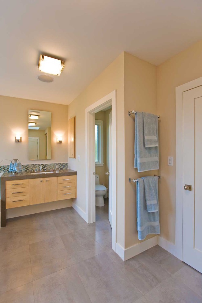 Cette photo montre une très grande salle de bain tendance en bois clair avec un lavabo encastré, un placard à porte plane, un plan de toilette en surface solide, une baignoire posée, une douche d'angle, WC à poser, un carrelage gris, des carreaux de porcelaine, un mur beige, un sol en carrelage de porcelaine et des toilettes cachées.