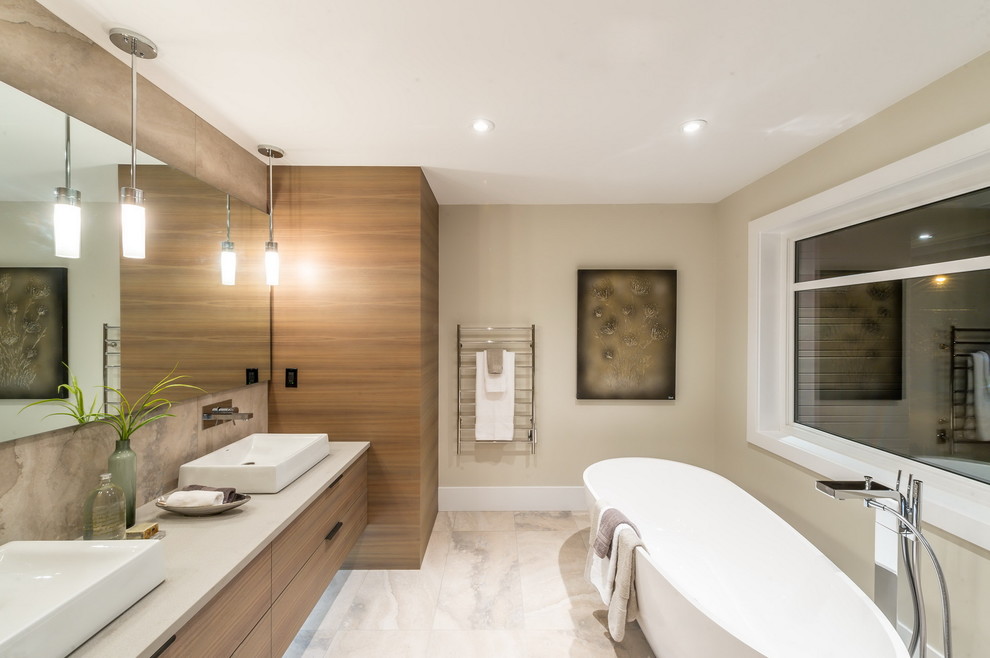 На фото: большая главная ванная комната в современном стиле с плоскими фасадами, светлыми деревянными фасадами, отдельно стоящей ванной, бежевой плиткой, каменной плиткой, мраморным полом, настольной раковиной и столешницей из искусственного камня
